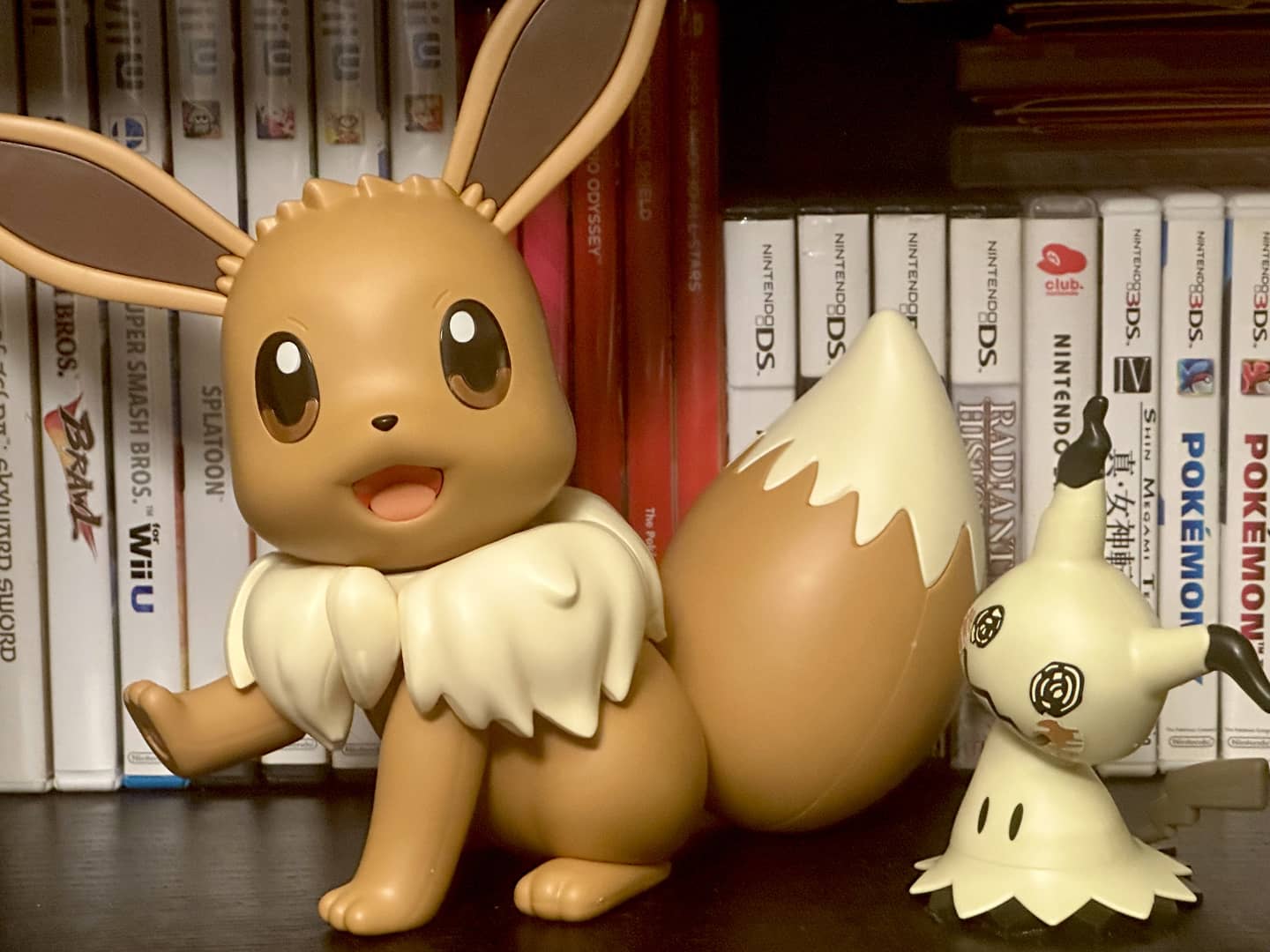 On the left, an Eevee standing with
its left paw raised. Its body is turned to the left, with its head facing the
camera. On the right is a Mimikyu, which is substantially smaller. Behind them
are several video game boxes for Nintendo consoles. The Eevee is about as high
as the Wii and Wii U DVD boxes, and the Mimikyu is a little shorter than the DS
boxes.